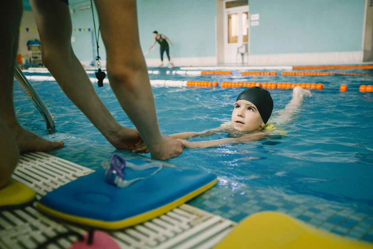 Young child in a black swim cap practicing swimming while an adult at the pool edge holds their hands; child is prone in the water and looking up toward the adult. Indoor 25 metre pool with orange lane ropes, tiled deck with a blue kickboard and goggles visible. Tone is calm, focused, and supported.